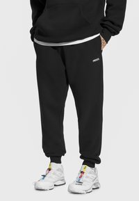 Czarne spodnie dresowe z elastycznymi mankietami, z logo po boku. Do tego białe buty sportowe z kolorowymi akcentami i sznurowadłami.