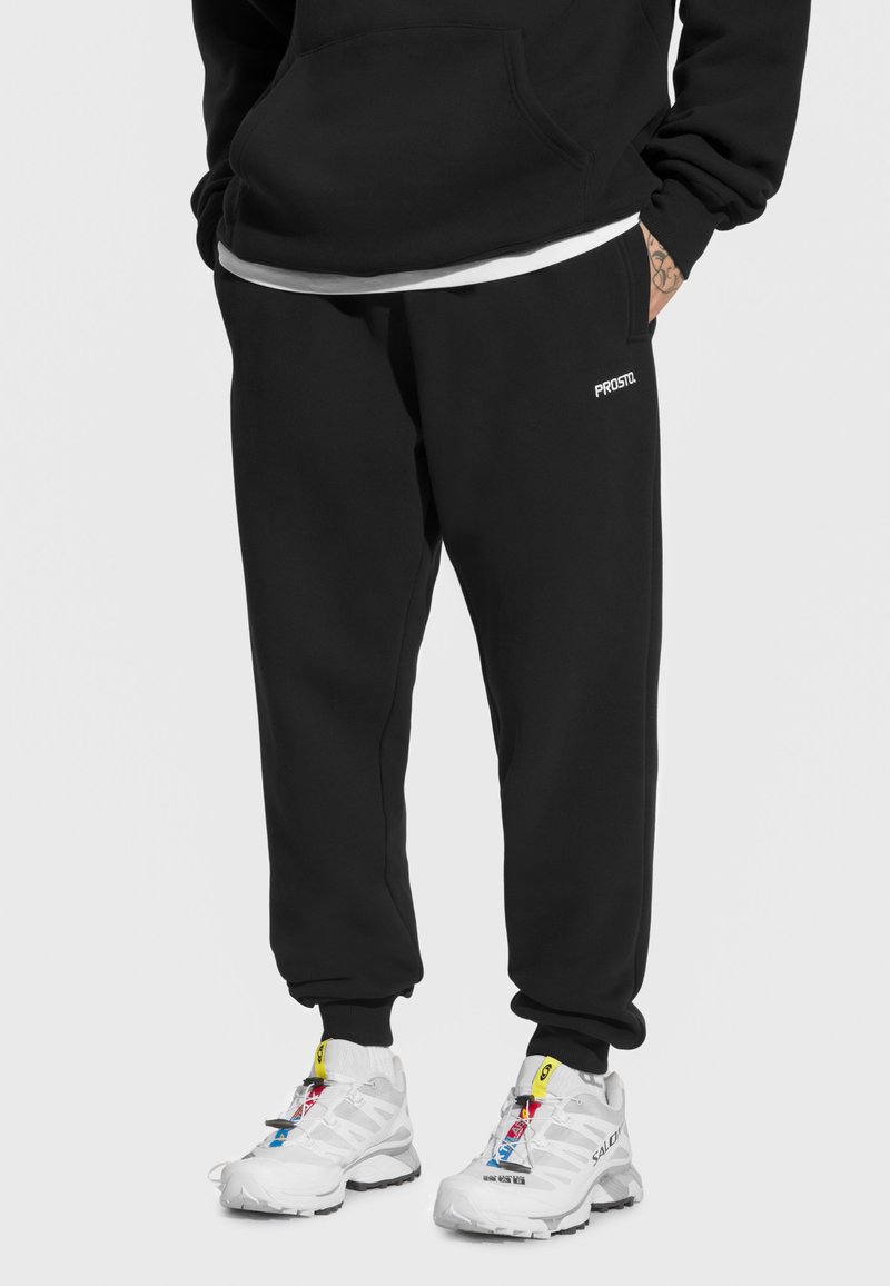 Czarne spodnie dresowe z elastycznymi mankietami, z logo po boku. Do tego białe buty sportowe z kolorowymi akcentami i sznurowadłami.