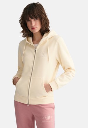 Hellgelbe Zip-Hoodie mit Kordelzügen, Vordertaschen und gerippten Bündchen. Getragen mit pinkfarbenen Jogginghosen, die mit weißem Logo-Druck versehen sind.