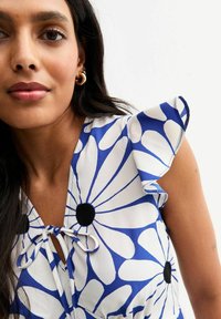 Flerfärgad blus med blå och vita blommönster, volangärmar, knyt detaljer vid halsen och svarta cirkelaccenter.