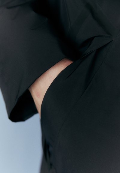 Sort beklædning med en glat, mat tekstur har et ovalt udsnit, der afslører hud. Designet har bløde draperinger omkring kanterne.