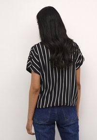 Femme aux cheveux longs et ondulés portant un chemisier à manches courtes à rayures verticales noires et blanches et un jean bleu, faisant face à un mur blanc.