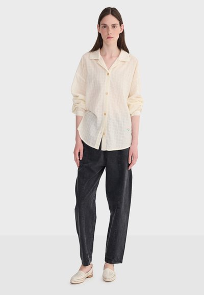 Jeune femme debout portant une chemise crème ample à boutons, un pantalon noir à jambes larges et des chaussures légères beige clair sans lacets, sur un fond uni.