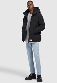 Schwarze Steppjacke mit Kapuze, ausgestattet mit einem Reißverschluss, quchiline Design und Fronttaschen, kombiniert mit hellblauen Jeans und schwarzen Stiefeln.