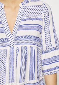 Blouse blanche avec des motifs géométriques et rayures bleues, un col en V profond et des manches trois-quarts larges avec des détails froncés.