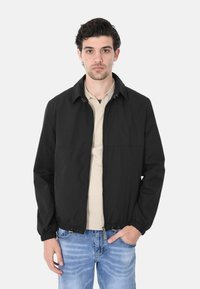 Giacca leggera nera con colletto alzato, polsini elasticizzati e chiusura con zip. Abbinata a una polo beige e jeans in denim.