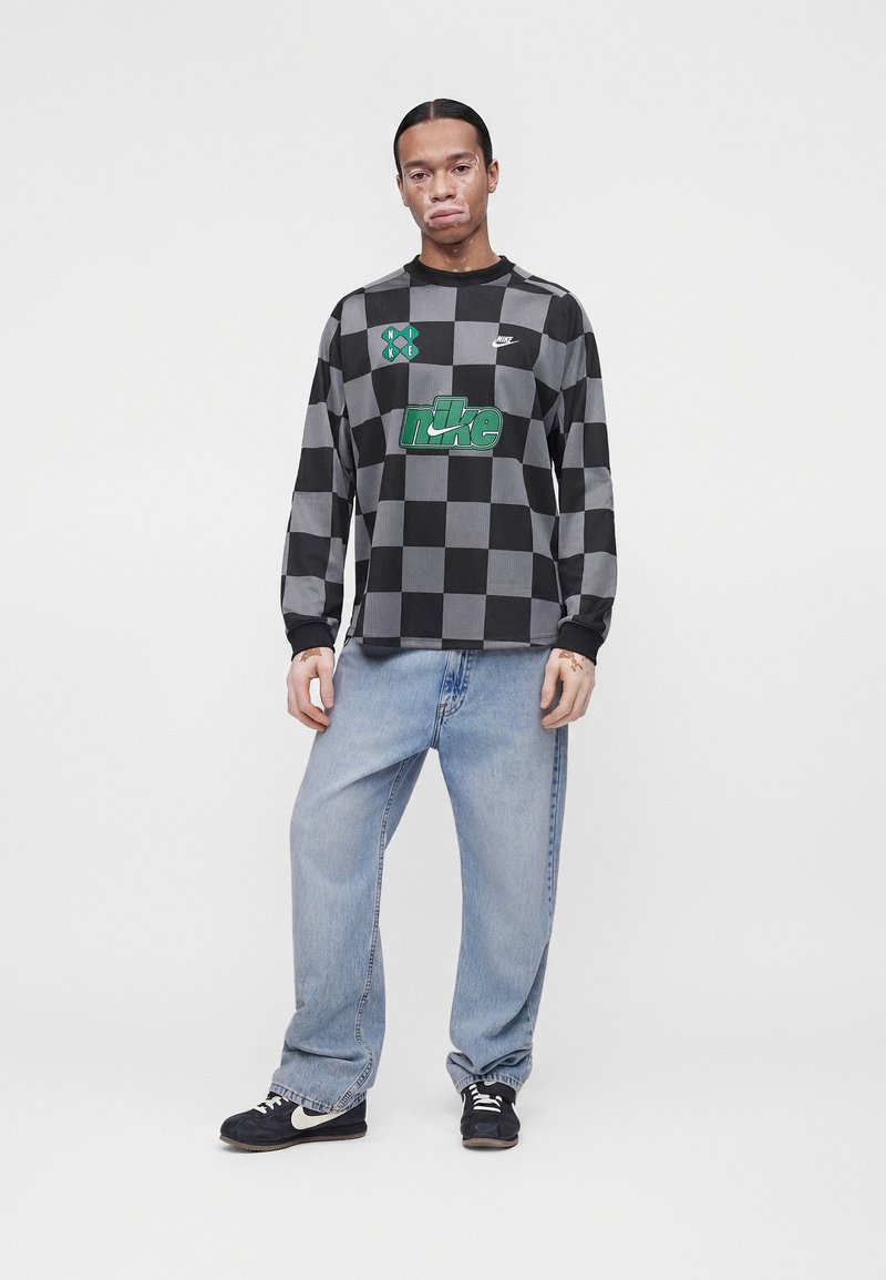 Mann mit Vitiligo, der ein schwarz-grau kariertes langärmeliges Nike-Shirt, hellblaue Jeans und schwarze Nike-Sneakers trägt, steht vor einem einfarbigen Hintergrund.