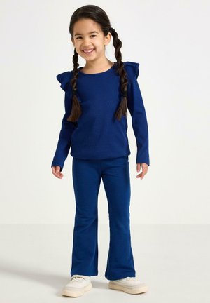 Laivastonsininen pitkähihainen paita, jossa on röyhelökoristeiset hartiat, yhdistetty sopiviin liehuviin housuihin. Asun täydentävät valkoiset tennarit.