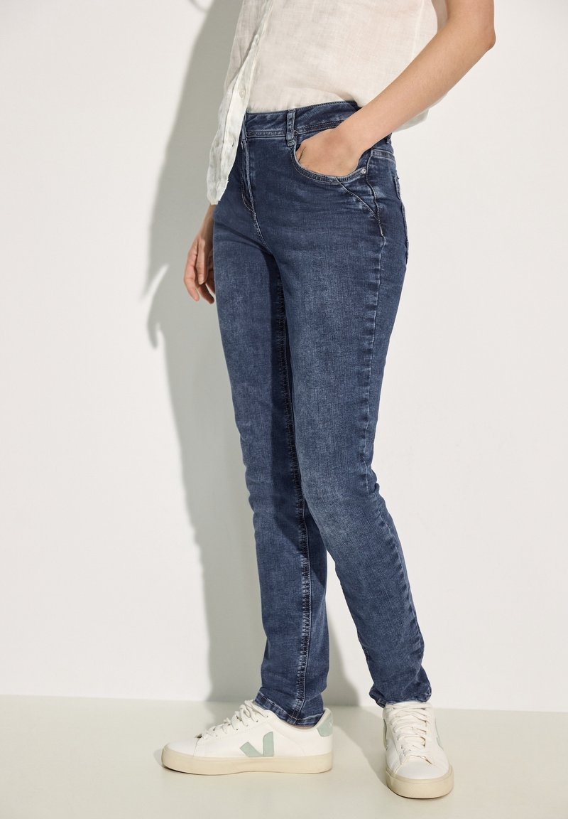 Skinny jeans in dark blue denim, featuring a mid-rise waist, front pockets, and a close fit. Paired with white sneakers with mint accents.