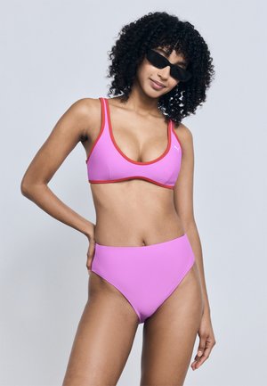 Femme aux cheveux bouclés portant des lunettes de soleil noires et un bikini violet bordé de rouge, debout devant un fond uni gris clair.