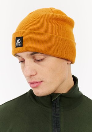 Oranje geribde beanie met een zwart logo patch, gedragen met een groene fleece met rits. Heeft een snug fit en een zachte textuur.