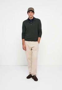 Zelený sveter so zipsom, námornícka polokošeľa, béžové nohavice a hnedé topánky. Model nosí károvanú čiapku. Jednoduchý dizajn, textúrovaná tkanina.