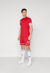 Rode sporttrui met witte strepen op de mouwen, bijpassende rode shorts, witte sokken met blauwe strepen en witte voetbalschoenen. Adidas-logo zichtbaar.