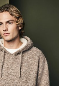 Brun strikket hoodie med en blød, cremefarvet fleece-foring rundt om halsen, med snøre i hætten og en afslappet pasform. Struktureret stof synligt.