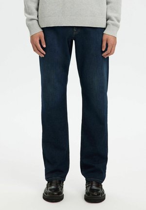 Personne portant un jean droit bleu foncé, des chaussures en cuir noires et un pull en maille gris clair, debout devant un fond blanc.