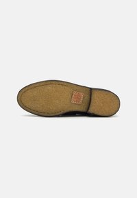 Suela de zapato con una base de goma texturizada marrón, que presenta una capa suave y peluda y un pequeño parche de cuero cerca del talón.