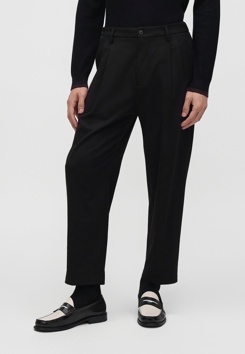 Personne portant un pantalon noir fuselé, des chaussettes noires et des mocassins noirs et blancs, debout devant un fond clair uni.