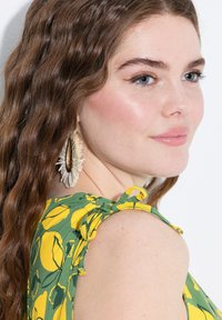 Jeune femme aux cheveux châtains ondulés portant de grandes boucles d'oreilles décoratives et un haut sans manches vert avec des motifs de feuilles jaunes.