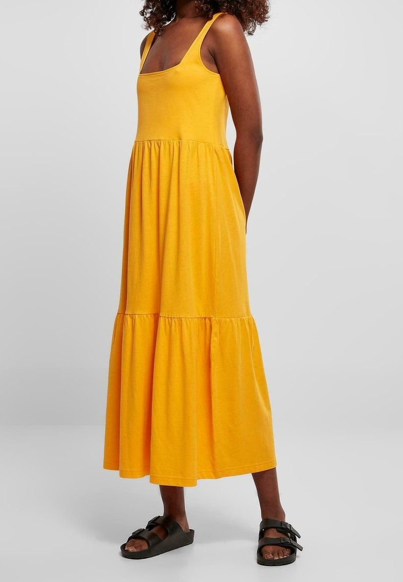 Femme portant une robe midi sans manches jaune à volants, associée à des sandales noires à boucle, debout devant un fond clair uni.
