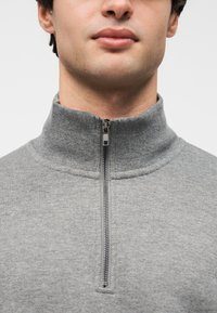 Γκρι φλις πουλόβερ με φερμουάρ και ψηλό γιακά, με ασημένιο φερμουάρ και ριμπ υφή στο λαιμό. Κοντινή άποψη του πάνω μέρους του κορμού.