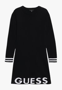 Musta neulemekko pitkähihaisilla ja pyöreällä kaula-aukolla. Ominaisuuksiin kuuluu valkoiset raidalliset yksityiskohdat hihoissa ja "GUESS"-logo helmassa.