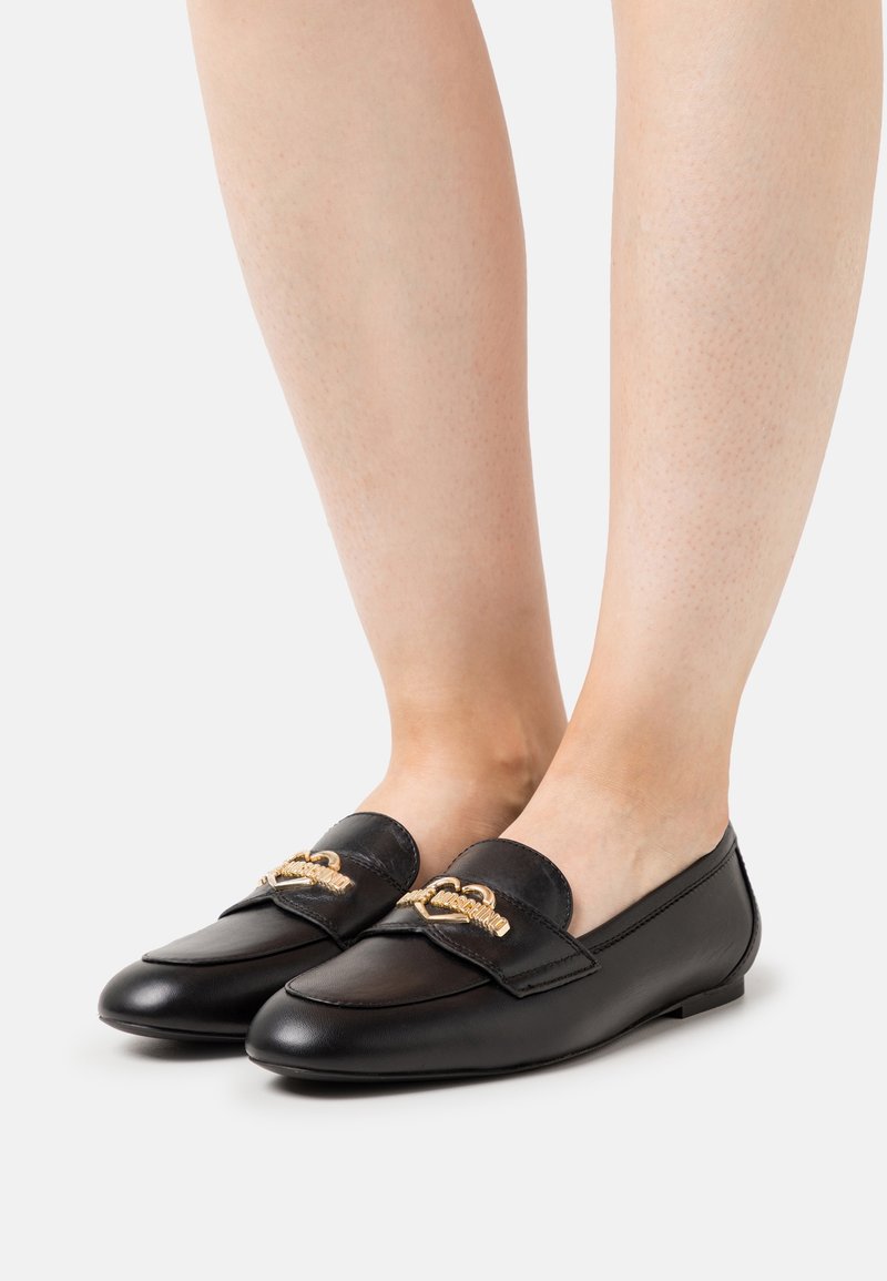 Mocassins en cuir noir avec détail chaîne dorée portés pieds nus, visibles de face sur fond blanc.