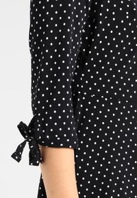 Černé šaty s bílými puntíky, mírně ohrnutými rukávy se stužkami na mašli, jemnou texturou látky a přiléhavým střihem.