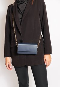 Borsa a tracolla in pelle blu navy con patta pieghevole, tracolla in catena dorata e texture liscia, indossata con un blazer nero e pantaloni.