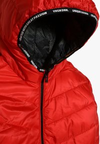 Chaqueta acolchada roja con cremallera negra y capucha forrada de tela negra, con un ribete de texto en blanco y naranja.