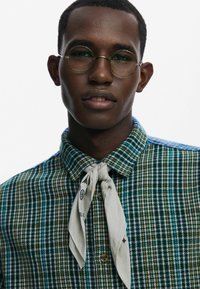 Koszula w kratę w odcieniach niebieskiego, zielonego i żółtego z kołnierzykiem zapinanym na guziki; z przodu lekko szary szalik związany wokół szyi z nadrukowanymi detalami.