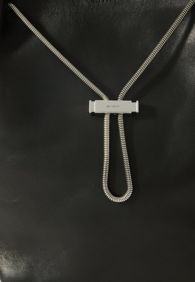 Superficie in pelle nera con una catena a serpente d'argento che forma un anello, tenuta da una chiusura metallica rettangolare incisa con "BY FAR."