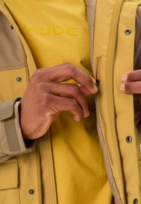Giacca trapuntata gialla con esterno beige, caratterizzata da una tasca con zip e dettaglio con bottoni a pressione, indossata su una camicia gialla abbinata.