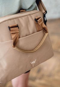 Tasche aus weichem, tanfarbigem Stoff, mit einer Vordertasche, kurzem Griff, verstellbarem Schulterriemen und einem weißen Logo. Strukturierten Oberfläche.