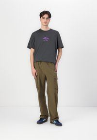 Gray cotton t-shirt with purple logo, paired with olive cargo pants featuring side pockets, and blue sneakers with three stripes.