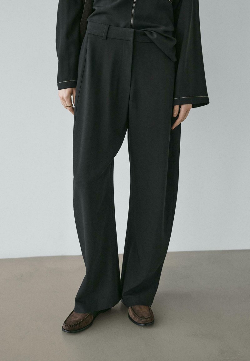 Black wide-leg trousers feature a smooth fabric, high waist, and a flat front design. Paired with brown loafers for contrast.