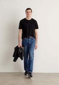 T-shirt noir, jeans en denim bleu, chaussures noires. Le modèle tient une veste en cuir noire. Style simple et décontracté avec des couleurs neutres.