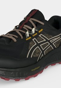 Μαύρο και καφέ παπούτσι τρεξίματος Asics με μπεζ κορδόνια, λευκές και κίτρινες λεπτομέρειες, και κόκκινη εξωτερική σόλα, με ενδιάμεση σόλα AmpliFoam.