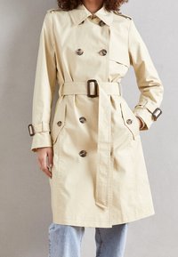 Beige värinen trenssitakki, jossa on kaksirivinen nappilista, suuret napit, vyötärökaitale ja sivutaskut. Takissa on terävä kaulus ja resorit hihansuissa.