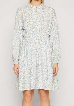 Femme portant une robe blanche à manches longues jusqu'aux genoux avec imprimé floral bleu clair et une petite fente à l'encolure devant.