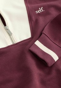 Felpa zip-up color marrone scuro con pannello crema, caratterizzata da "self." ricamato sul petto e polsini a righe a costine.