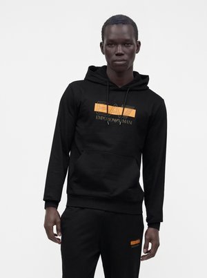 Mies käyttää mustaa Emporio Armani -hupparia ja siihen sopivia mustia housuja, joissa on oranssit logopatchit, seisoo yksiväristä valkoista taustaa vasten.