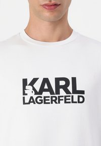 Hvit bomulls T-skjorte med en fremtredende svart trykt logo "KARL LAGERFELD" med et grafisk av en stilisert figur. Korte ermer, rund hals.