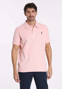 Polo-shirt rose clair en tissu lisse, avec un col classique et des manches courtes, arborant un petit logo noir sur la poitrine gauche.