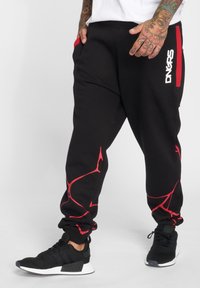 Black sweatpants with red accents, elastic waistband, and abstract red pattern. Paired with black athletic shoes featuring white soles.