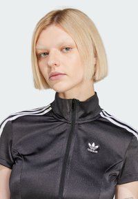 Zwarte sportjack met rits en korte mouwen, witte gestreepte accenten op de schouders en een klein wit Adidas-logo op de borst.