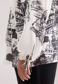 Blusa bianca con schizzi architettonici neri, maniche arricciate con nastri e una texture liscia. Abbinata a pantaloni neri.