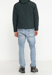 Chaqueta con capucha de color verde oscuro con un acabado texturizado, llevada sobre jeans azul claros y zapatillas negras con acentos amarillos. Vista trasera.