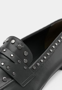 Mocassins en cuir noir avec des clous métalliques argentés le long de la sangle et des côtés, présentant une texture lisse et une silhouette classique.