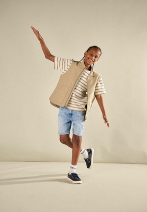 Child in beige vest, striped shirt, denim shorts, and sneakers balancing on one foot with arms extended against plain background.