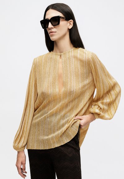 Γυναίκα που φοράει μεγάλα μαύρα γυαλιά ηλίου, μια χρυσή ριγέ μπλούζα με άνοιγμα στο λαιμό και μαύρο υφασμάτινο παντελόνι, ποζάροντας με το ένα χέρι στην τσέπη.